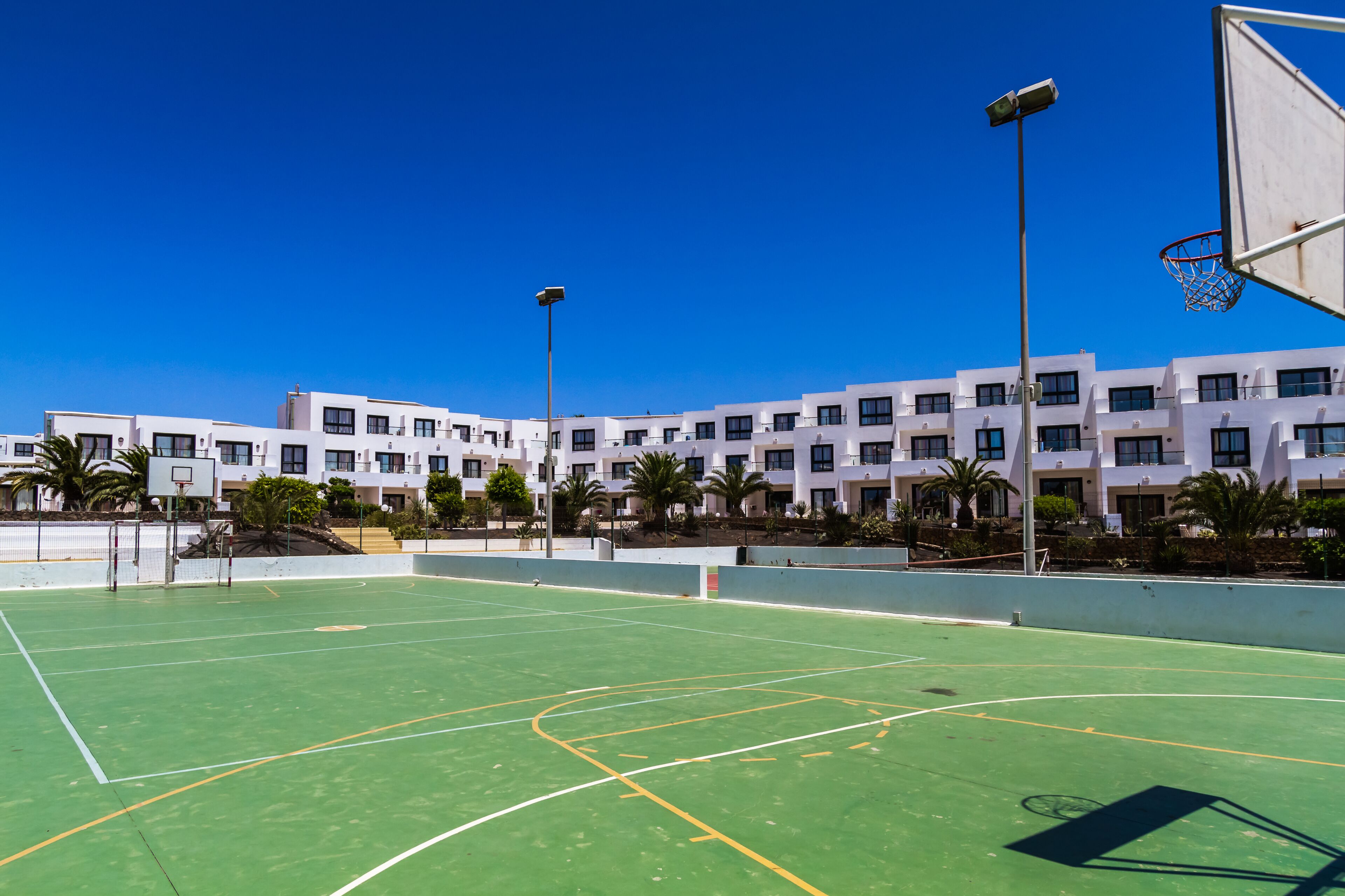 Una cancha de baloncesto al aire libre con un cielo azul despejado, rodeada de edificios blancos con múltiples ventanas y balcones, y palmeras.