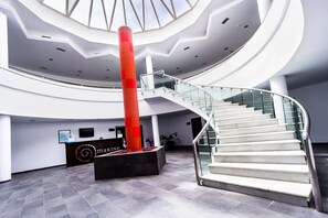 Interior moderno com escadaria central, balcão de recepção e uma grande janela de vidro.