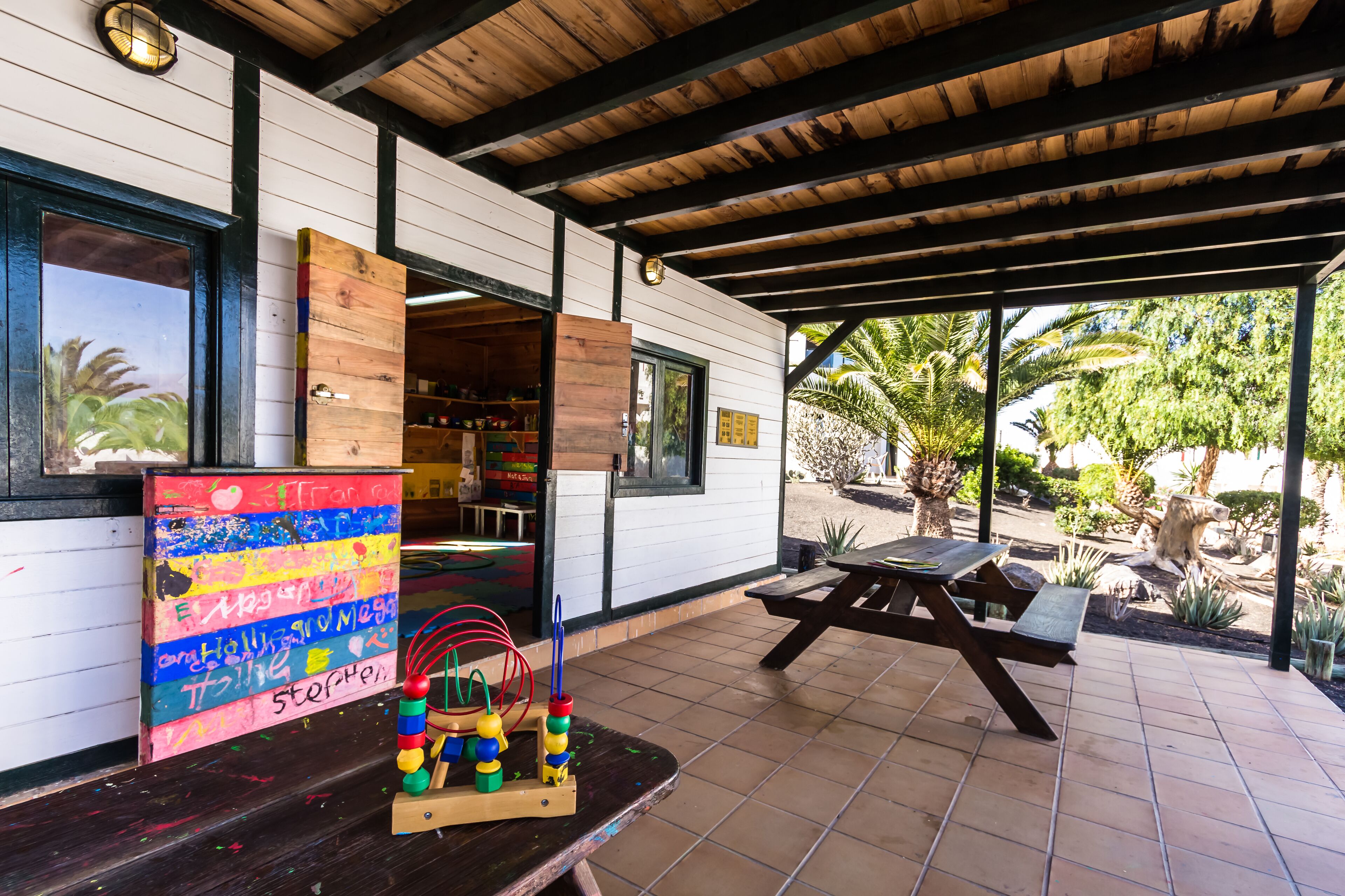 Ein Patio im Freien mit einem Holztisch und Bänken, einem farbenfrohen Bild an der Wand und Blick auf den umliegenden Garten.