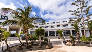 A modern building with a courtyard featuring palm trees, a small waterfall, and a seating area with steps leading up to the entrance. - Hotel BlueBay Lanzarote - All inclusive (Teguise)