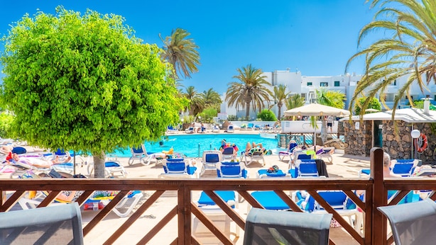A poolside area with lounge chairs, a swimming pool, and palm trees. - Hotel BlueBay Lanzarote - All inclusive (Teguise)