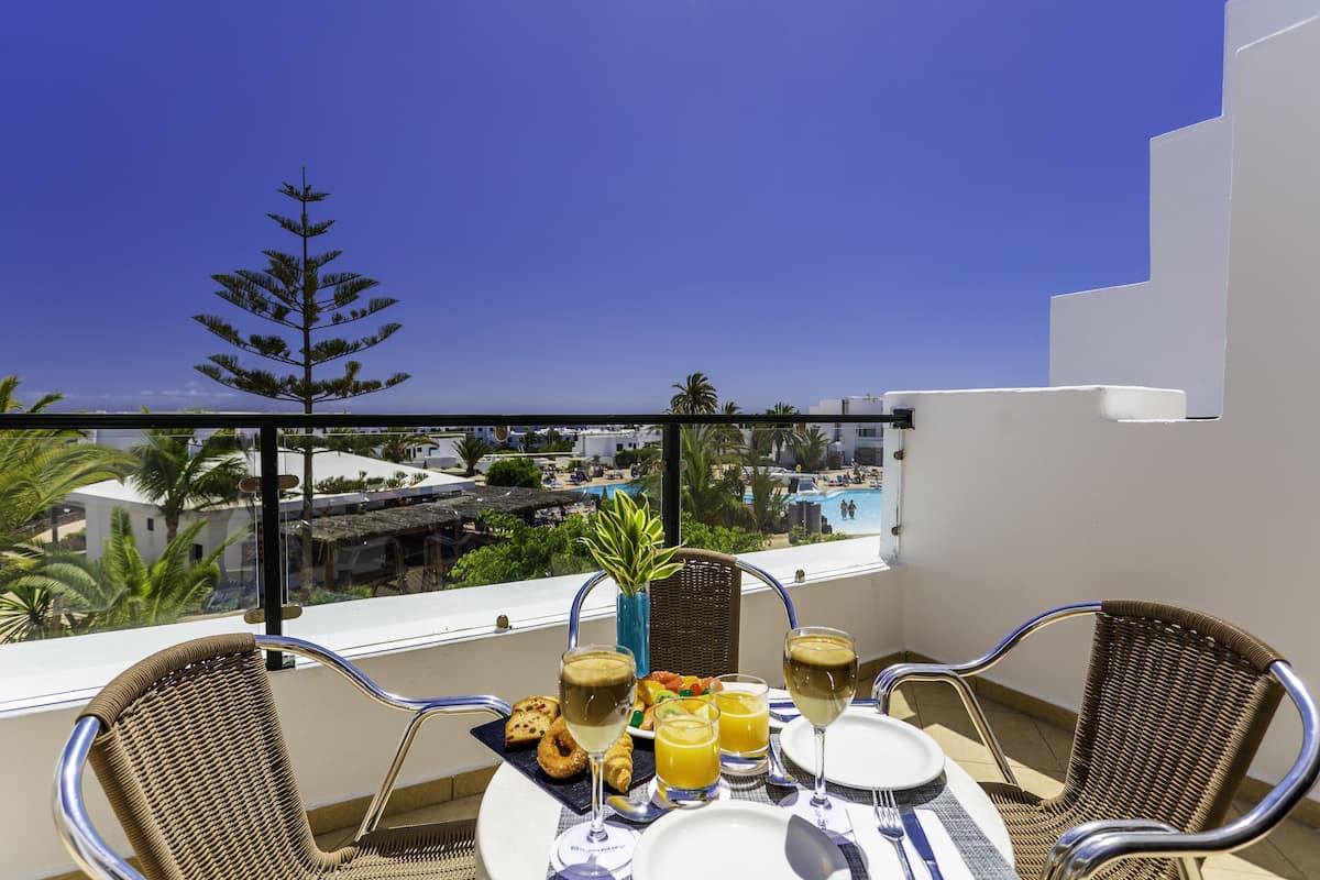A balcony with a table set for breakfast, including croissants, orange juice, and wine glasses, overlooking a pool and buildings.