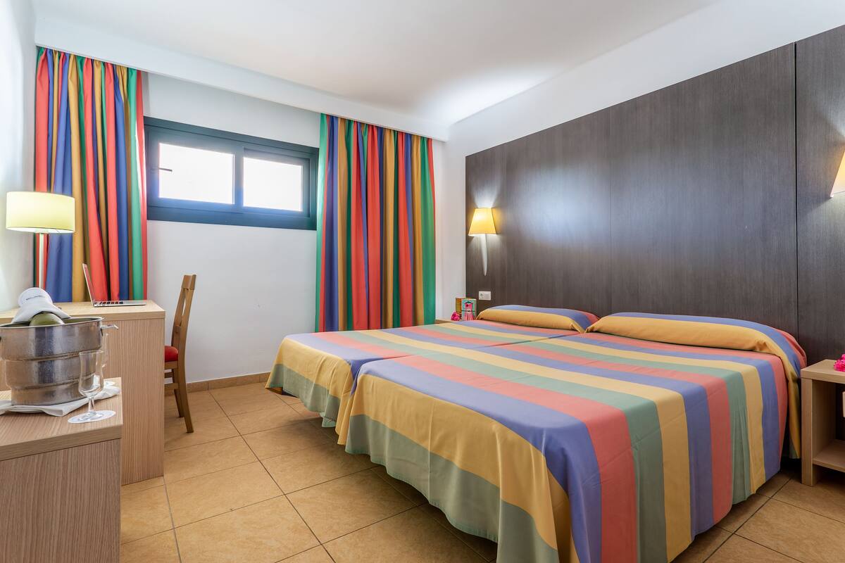 A hotel room with a bed, a desk, a chair, a lamp, and a window.
