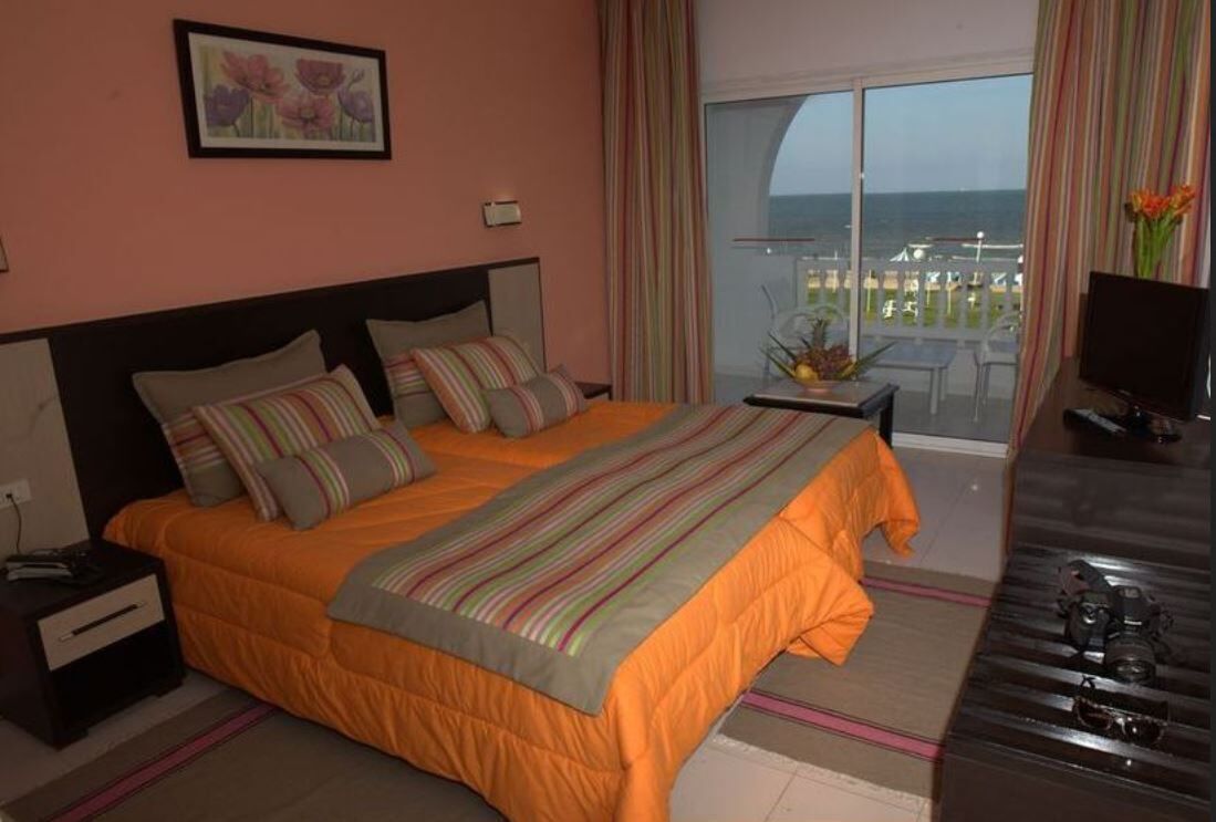Habitación de hotel con cama, mesita de noche, televisor, balcón con vista al mar y un cuadro en la pared.