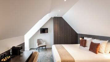 Ein modernes Hotelzimmer mit schrägverdeckter Decke, einem großen Bett mit vielen Kissen, einem Schreibtisch mit Stuhl und einem an der Wand montierten Fernseher.