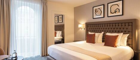 Habitación de hotel con una cama grande, una mesita pequeña y una silla. Se ve una ventana con cortinas, una lámpara de pared y cuadros sobre la cama.