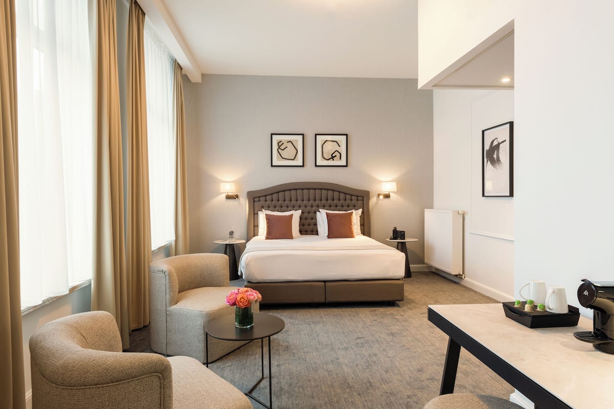 Ein Hotelzimmer mit einem großen Bett, zwei Nachttischen, einem kleinen Couchtisch, einer Couch und einem Schreibtisch. An der Wand hängen zwei gerahmte Kunstwerke und auf dem Boden steht eine Heizung.