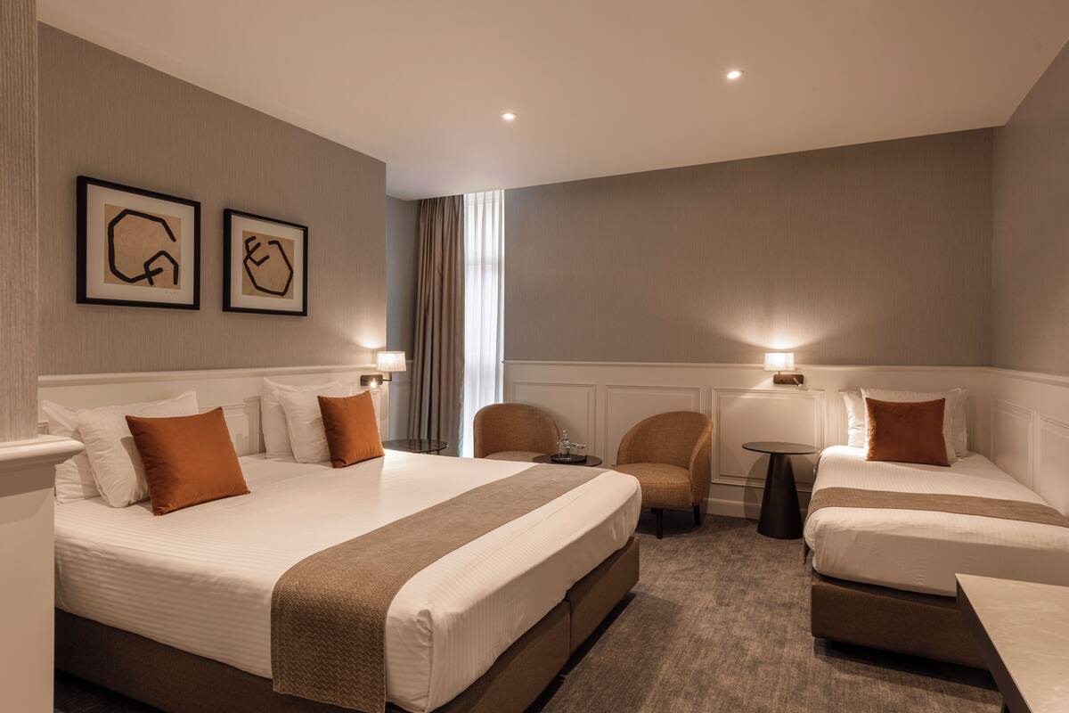 Ein Hotelzimmer mit zwei Betten, einer Sitzecke mit Sesseln, einem kleinen Tisch und gerahmten Kunstwerken an der Wand.