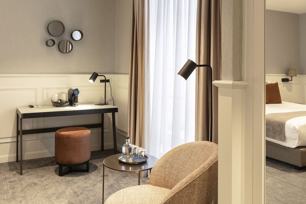 Ein Hotelzimmer mit Schreibtisch, Stuhl, rundem Hocker, einem kleinen Tisch mit Lampe und einem Bett mit Kissen.
