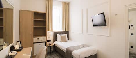 A hotel room with a bed, a desk with a chair, a small bedside table, a TV mounted on the wall, a wardrobe, and a window with curtains.