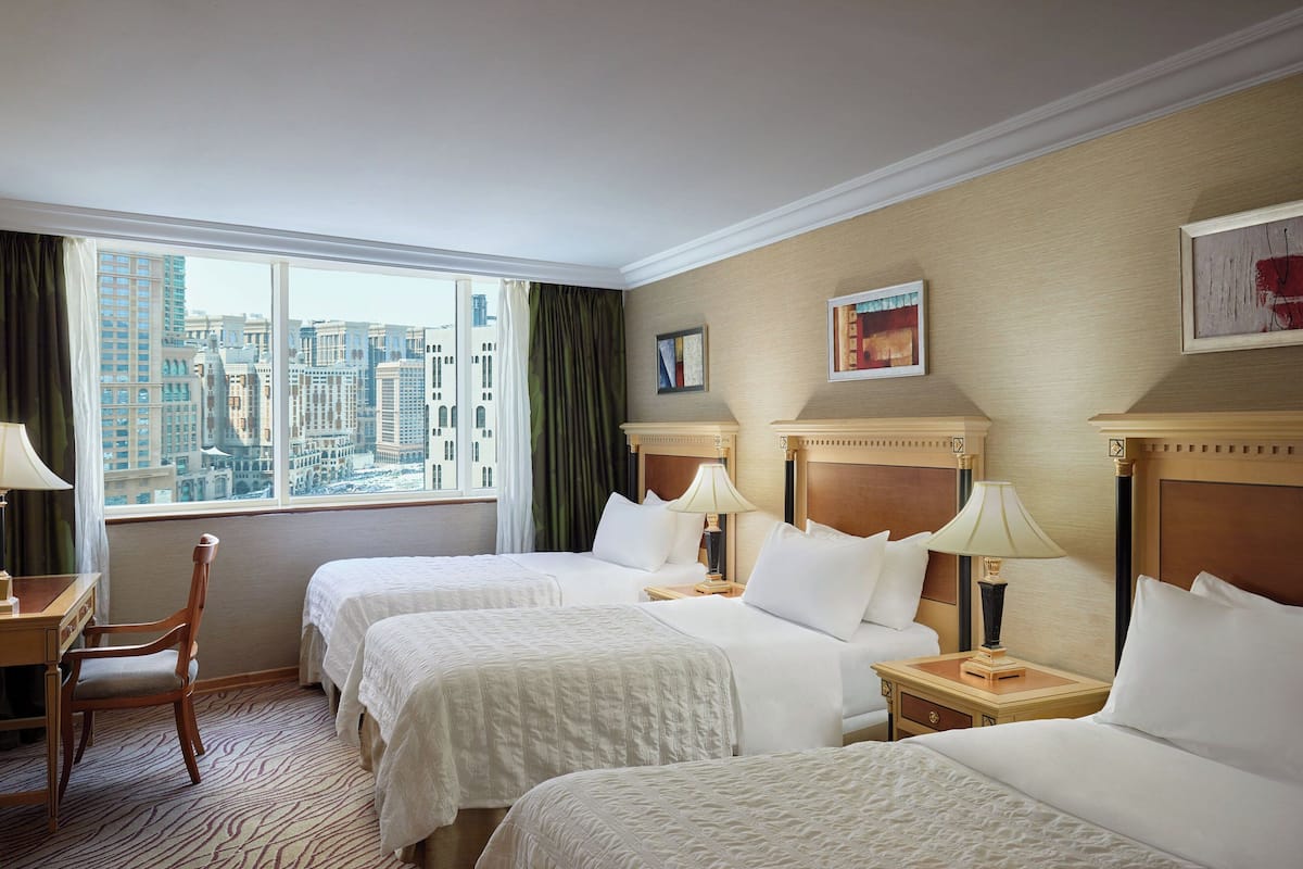 A hotel room with two beds, a desk, and a chair. There is a large window with a city view.