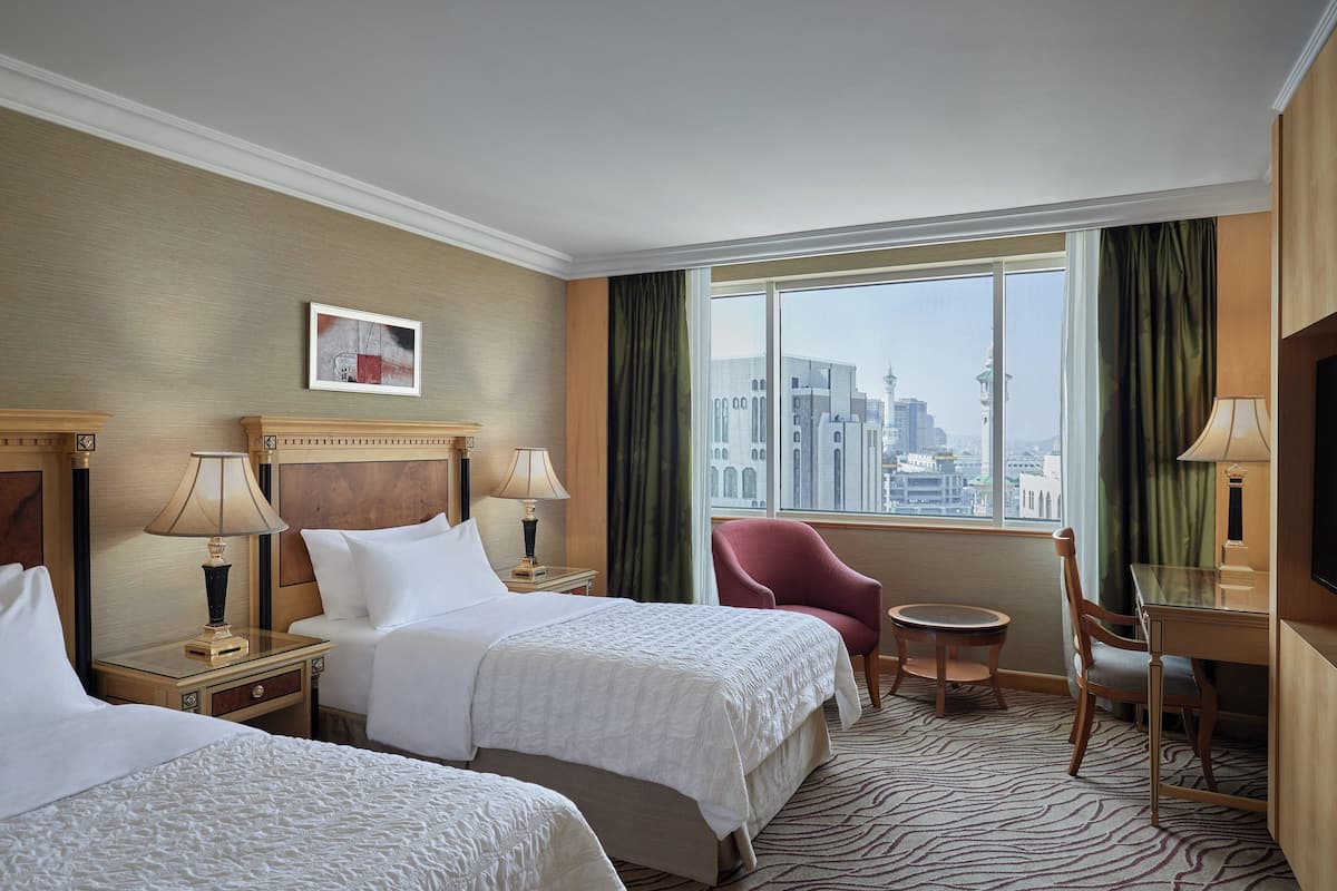 A hotel room with two beds, a desk, a chair, and a large window offering a city view.