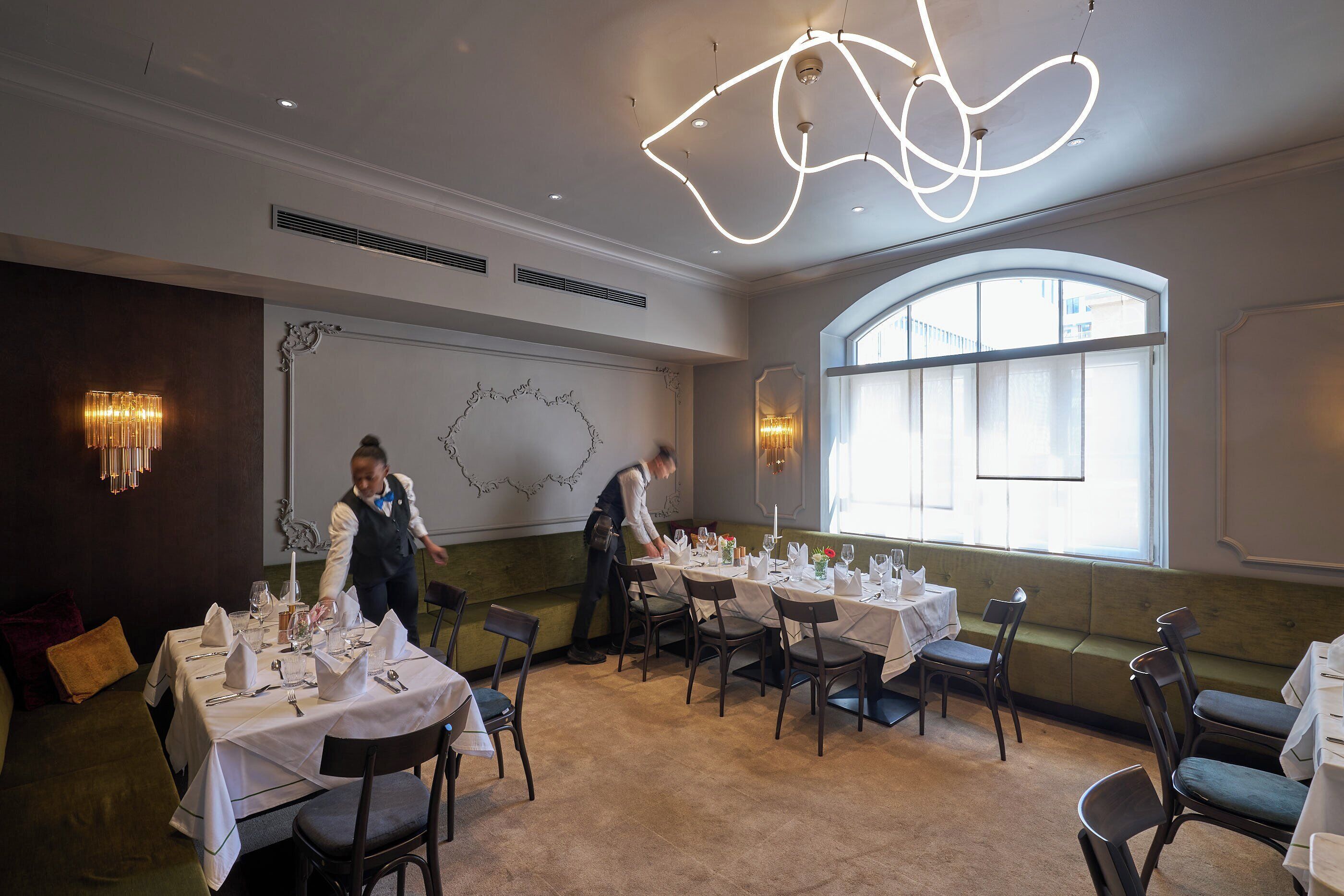 Un espace repas avec des tables dressées pour le service, une grande fenêtre en arc de cercle et un luminaire moderne.