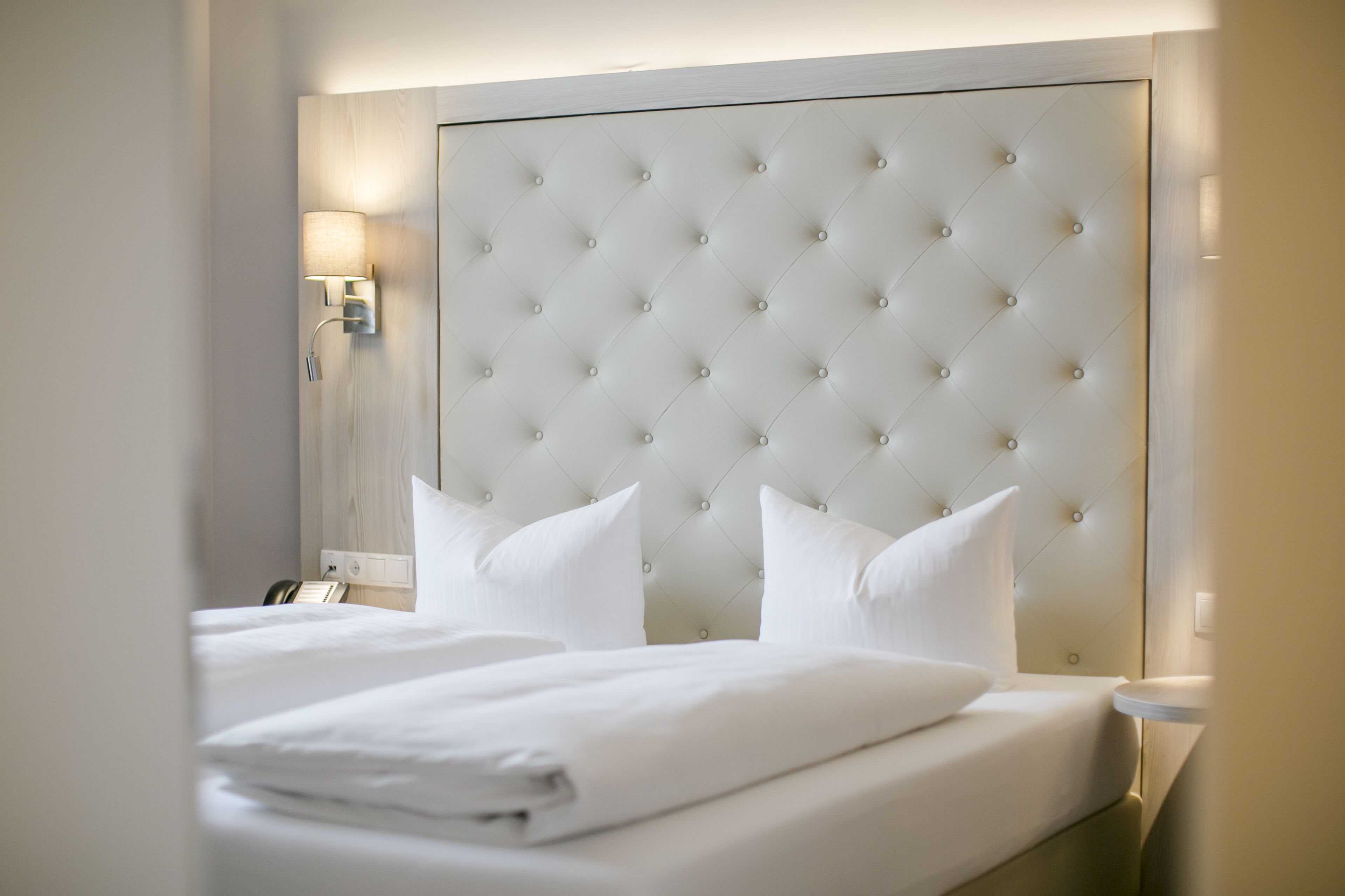A hotel room with a tufted headboard, two beds, white bedding, a bedside table, and a wall-mounted lamp.