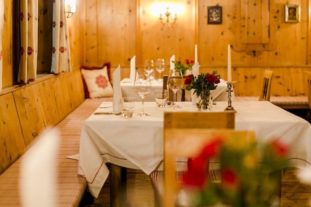 A dining table set with white tablecloth, candles, and glassware in a wooden room with a bench and floral curtains.