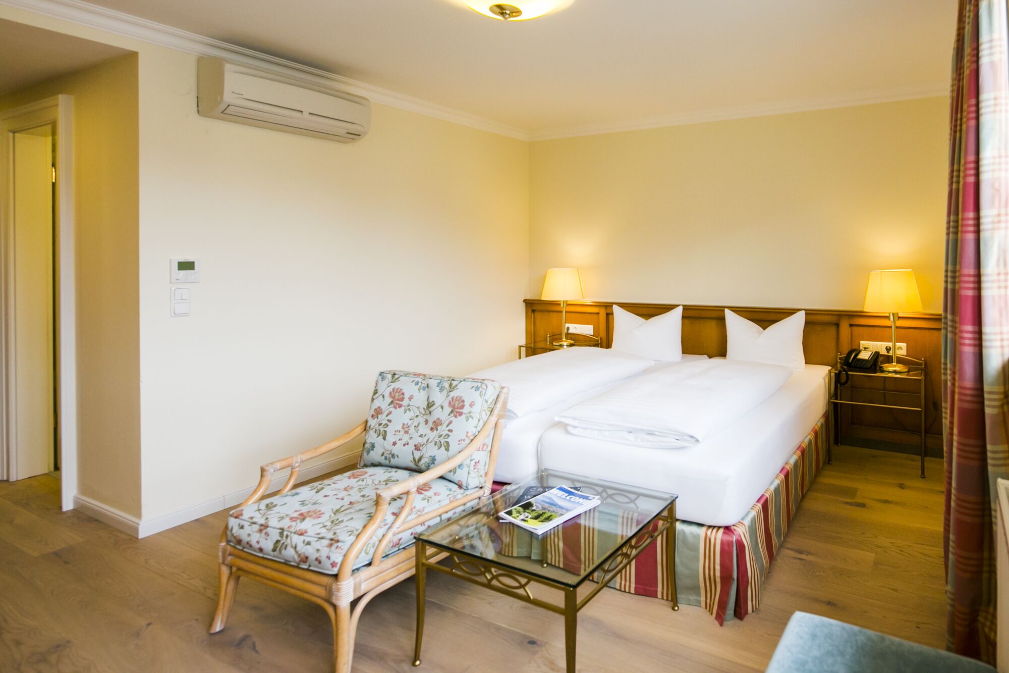 Quarto de hotel com cama, uma cadeira, uma mesinha e mesa de cabeceira com abajur.