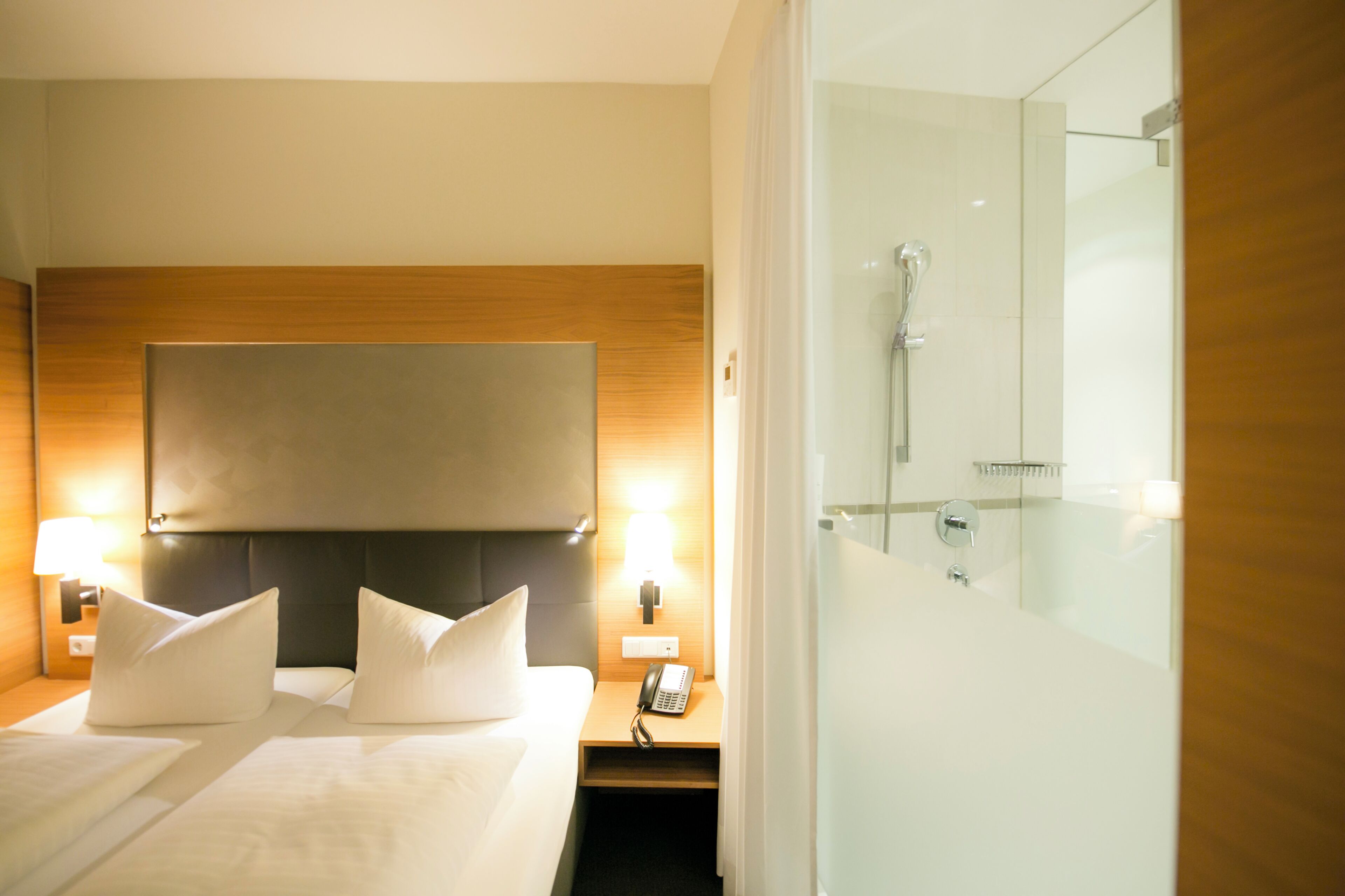 A hotel room with a bed, bedside lamps, a telephone, and a glass-enclosed shower.