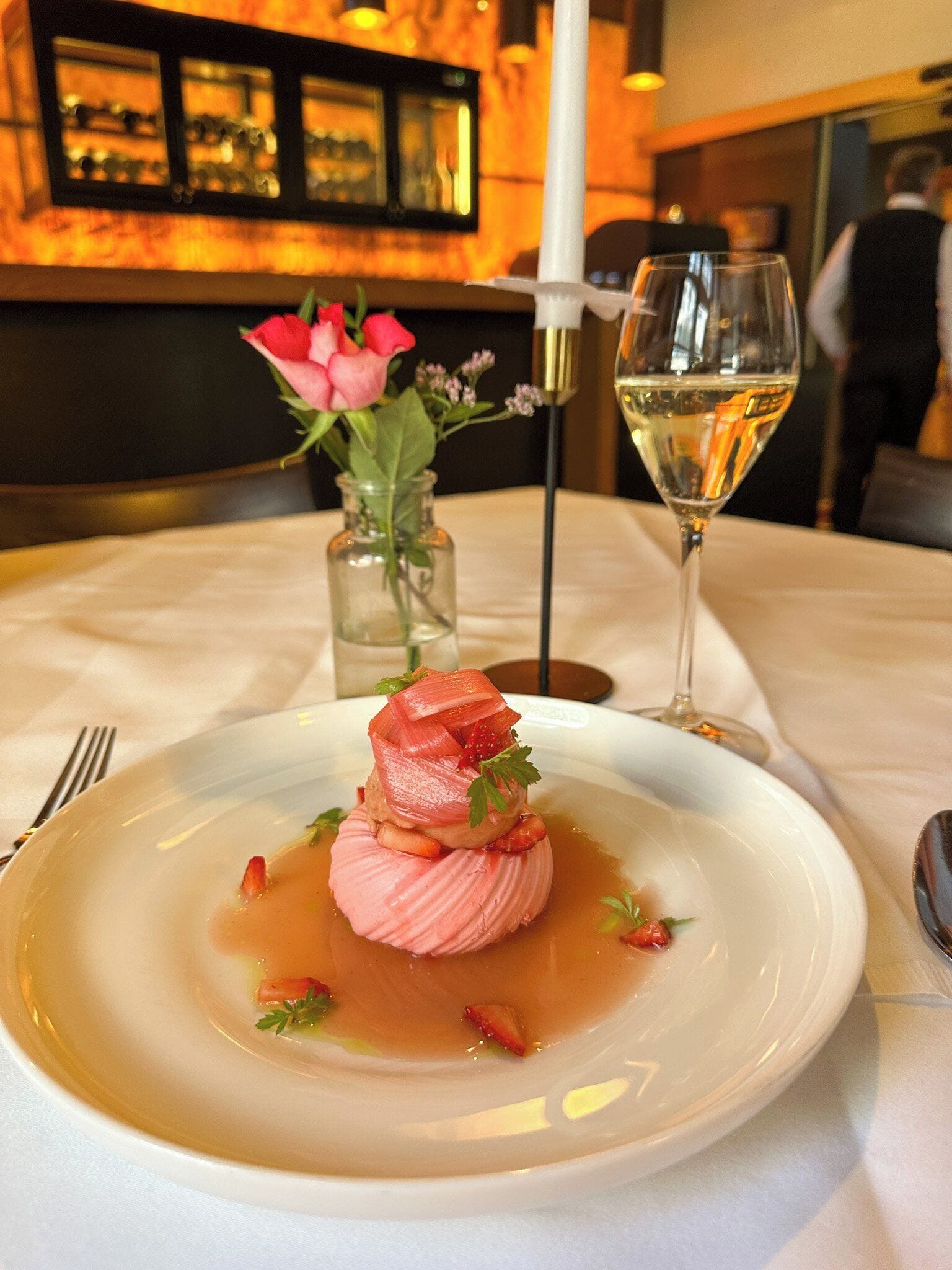 Dessert auf einem Teller mit einem Glas Weißwein und einer Vase mit Blumen auf einem Tisch.
