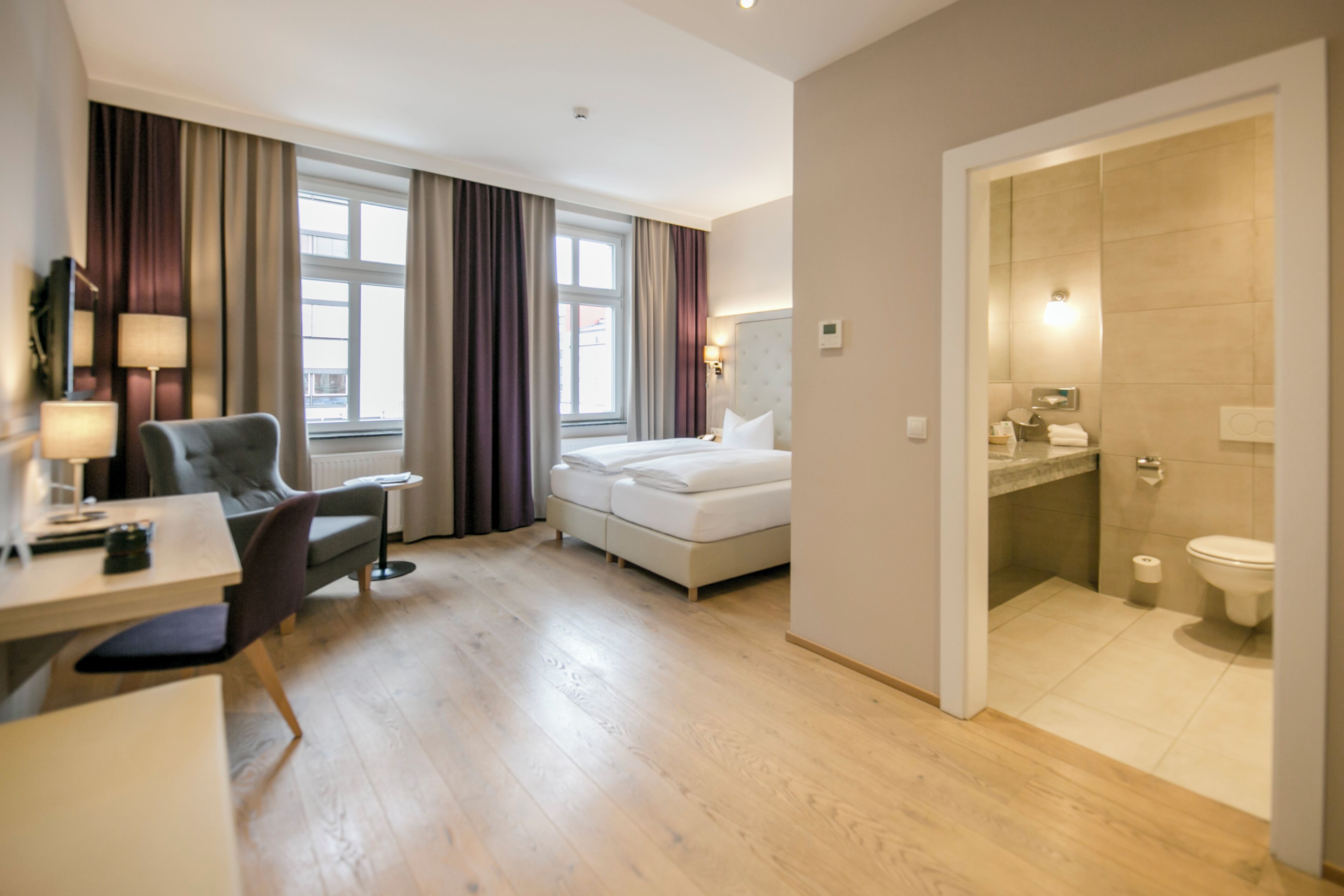 A hotel room with a bed, a desk with a chair, a bathroom with a toilet and sink, and a view of the outside through large windows.
