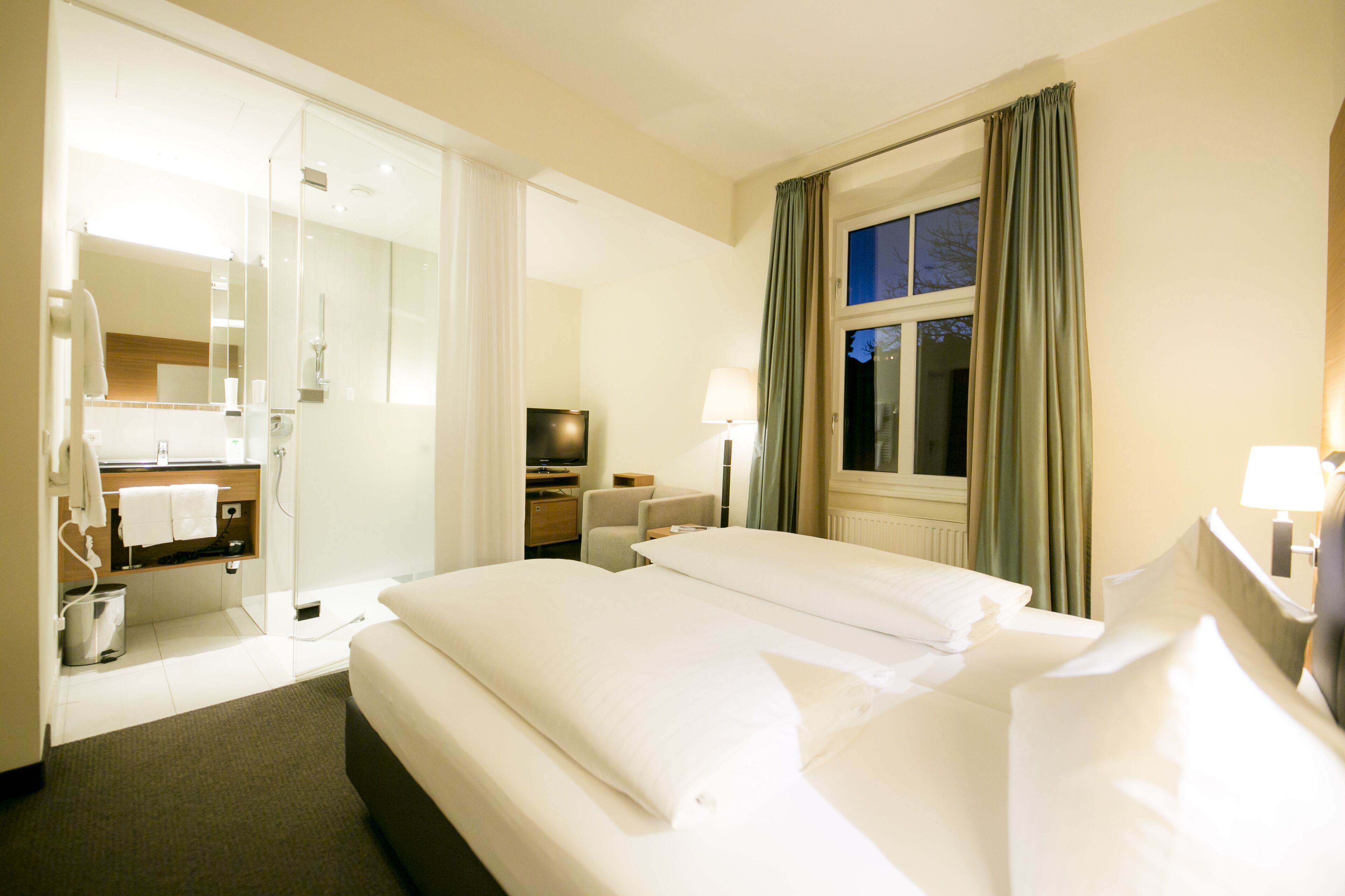 A hotel room with a large bed, a desk, a chair, a TV, and a bathroom with a mirror and towel rack.