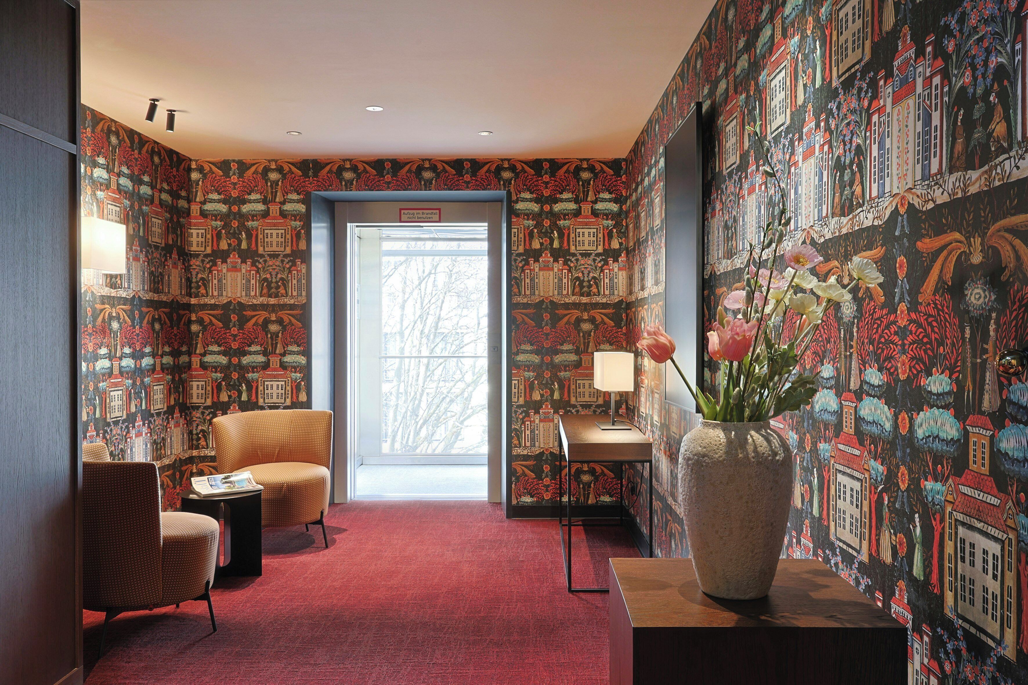 Ein Zimmer mit gemustertem Wandpapier, einem roten Teppich, einer großen Vase mit Blumen und zwei Stühlen.