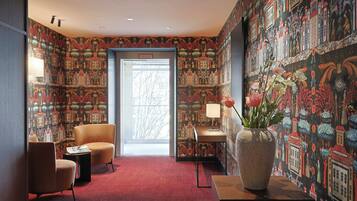 Ein Zimmer mit gemustertem Wandpapier, einem roten Teppich, einer großen Vase mit Blumen und zwei Stühlen.