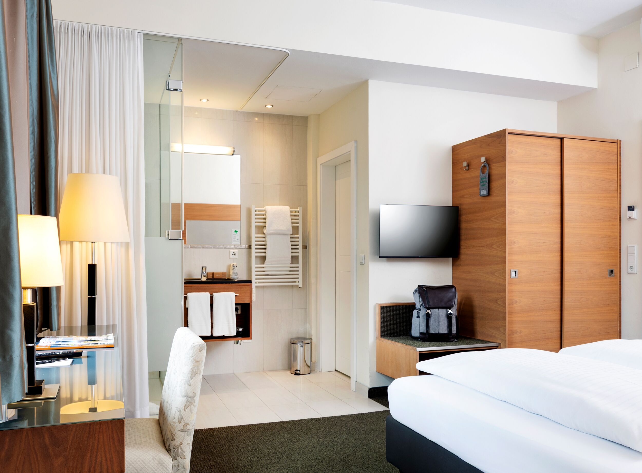 Habitación de hotel con cama, escritorio con lámpara, televisor, armario, espejo, toallero y basurero.