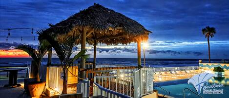 A beach resort with a thatched-roof structure, a pool area, and a view of the ocean at sunset.