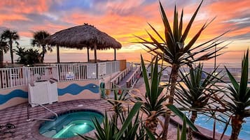 Área da piscina com guarda-sol de palha, palmeiras e vista para o pôr do sol no oceano.