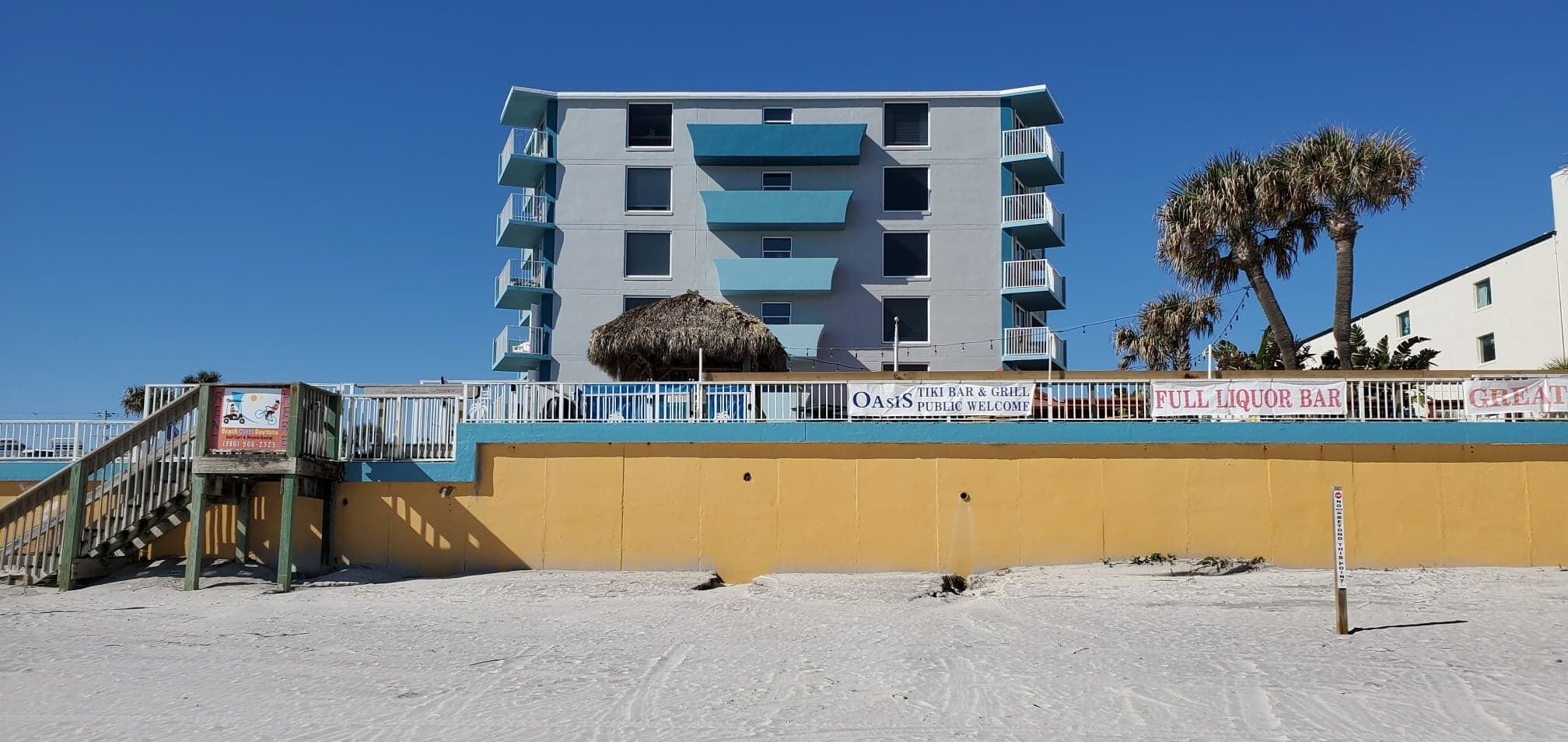 Un edificio de varios pisos con balcones, una playa al frente y palmeras.