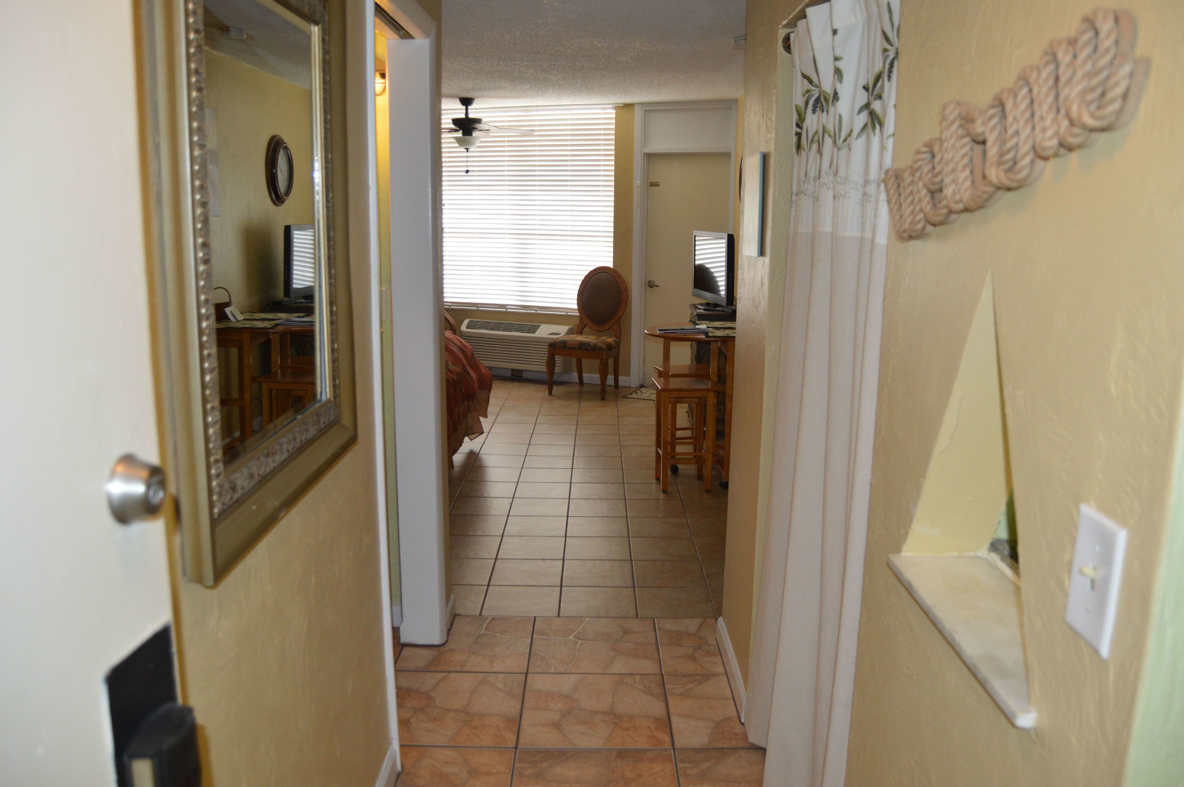 A hallway with a door leading to a room with a bed, a chair, a desk, and a window with blinds.