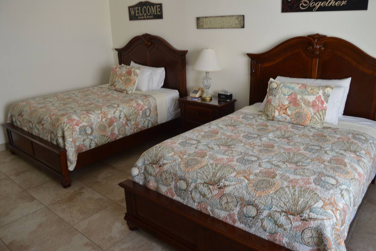 Two double beds with matching floral bedspreads, a nightstand with a lamp, and a wall sign that reads "WELCOME Family & Friends".