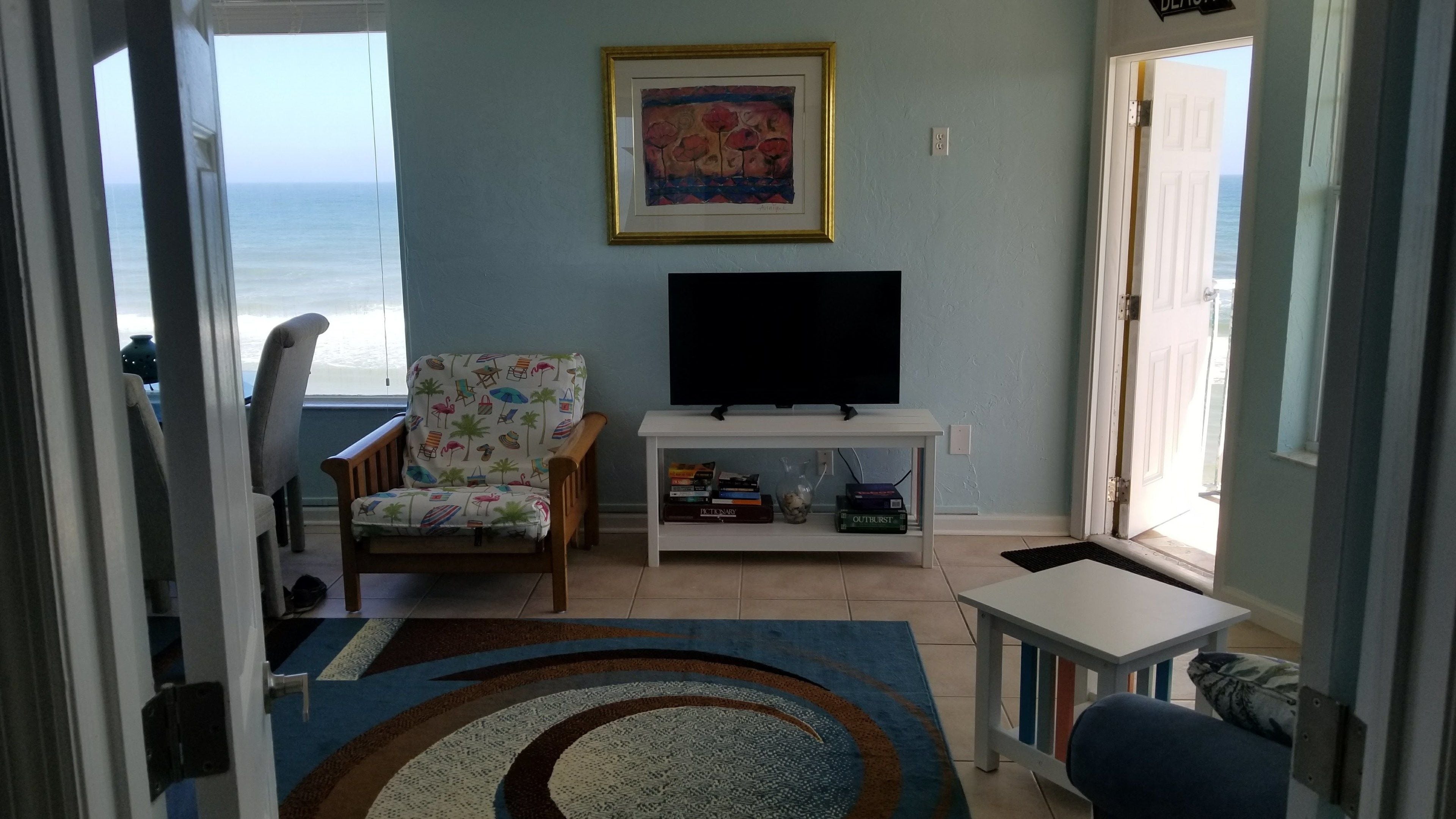 A living room with a beach view, a television, a chair with a tropical print, and a small table with books.