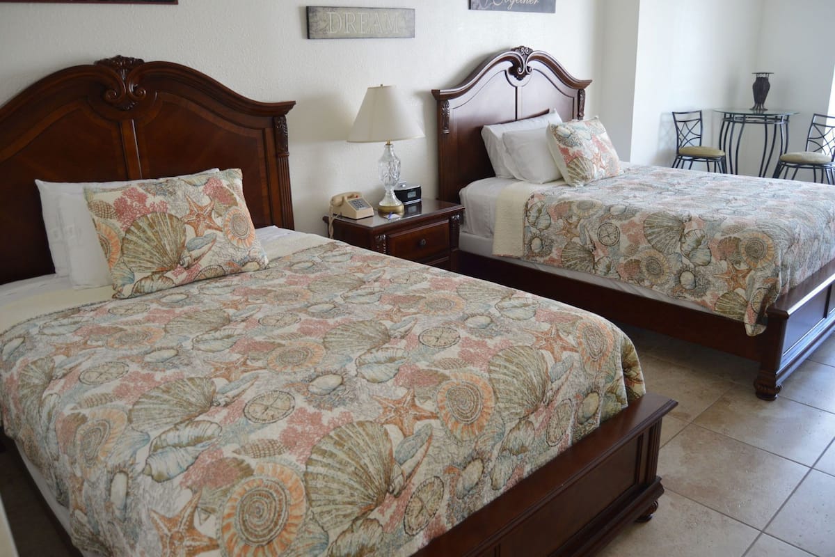 Two double beds with matching floral bedspreads, a bedside table with a lamp, and a small table with chairs in the corner.