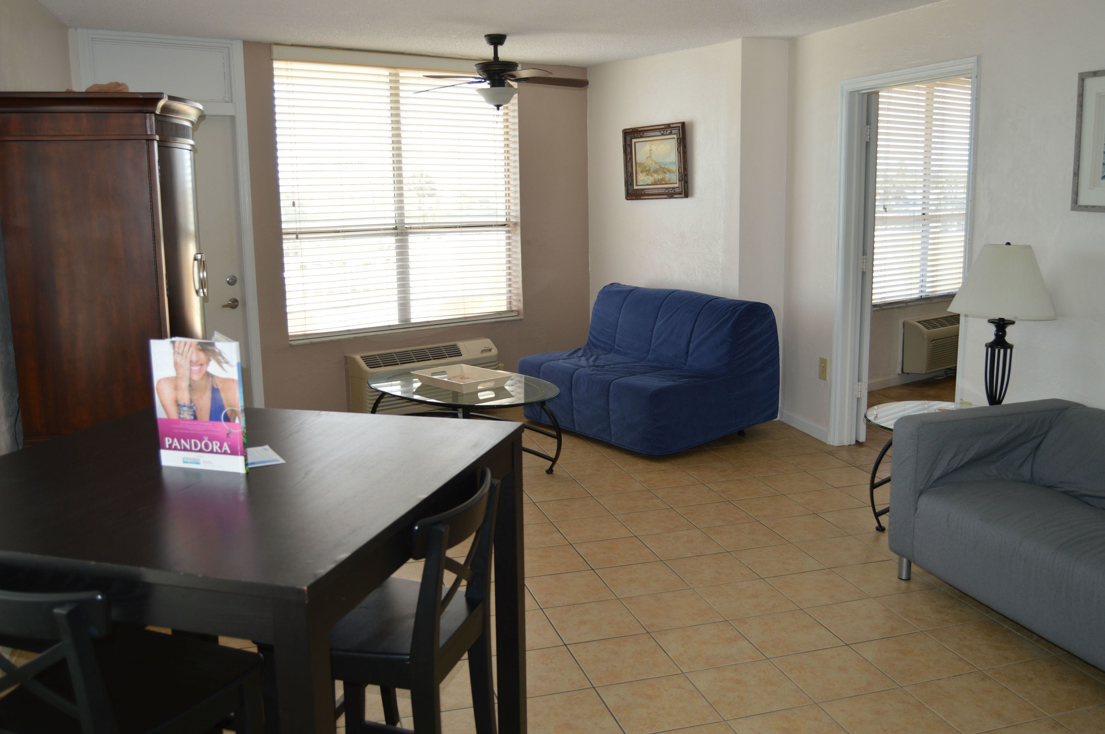 A living room with a dining table, a blue sofa, a grey sofa, a ceiling fan, and a window with blinds.