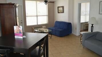 A living room with a dining table, a blue sofa, a grey sofa, a ceiling fan, and a window with blinds.