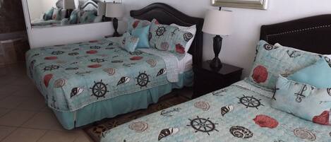 Un dormitorio con dos camas, cada una con una cubierta de edredón de temática náutica, una mesita de noche con lámpara, un espejo y un cuadro con una imagen de un velero en la pared.
