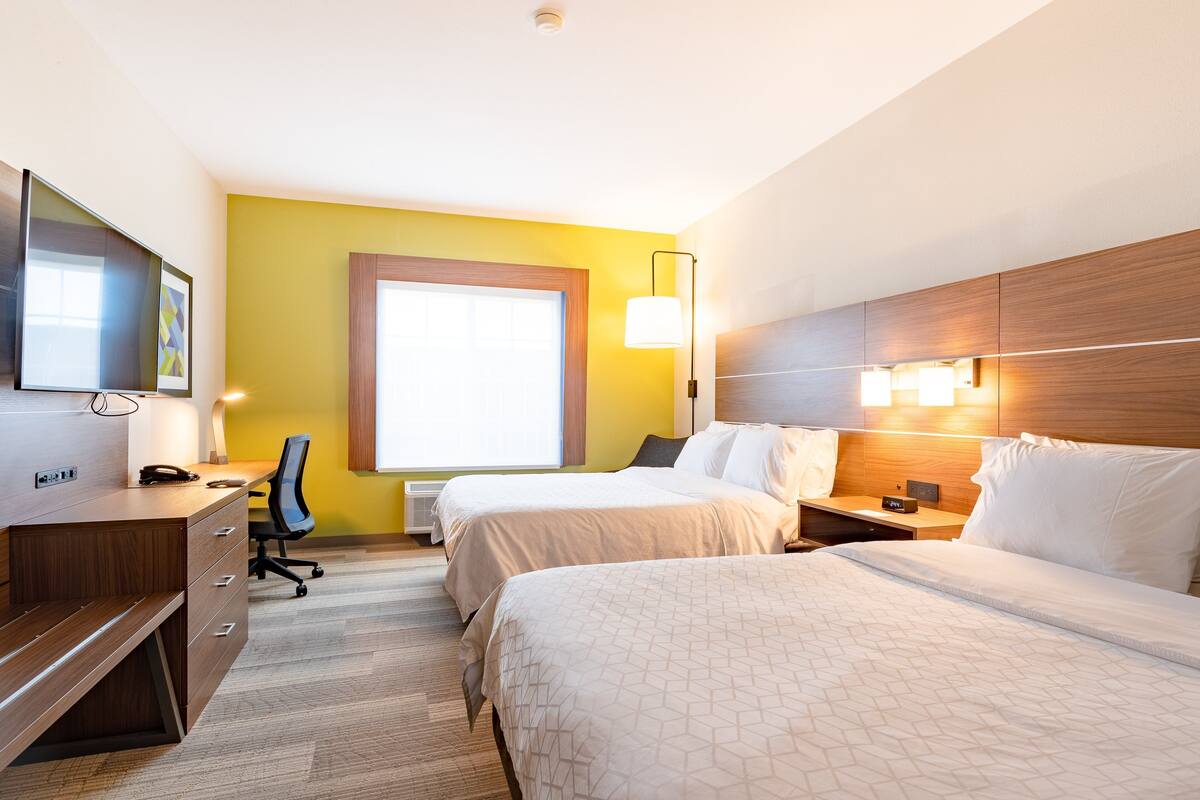 A hotel room with a wooden desk, two beds, a chair, and a large window.