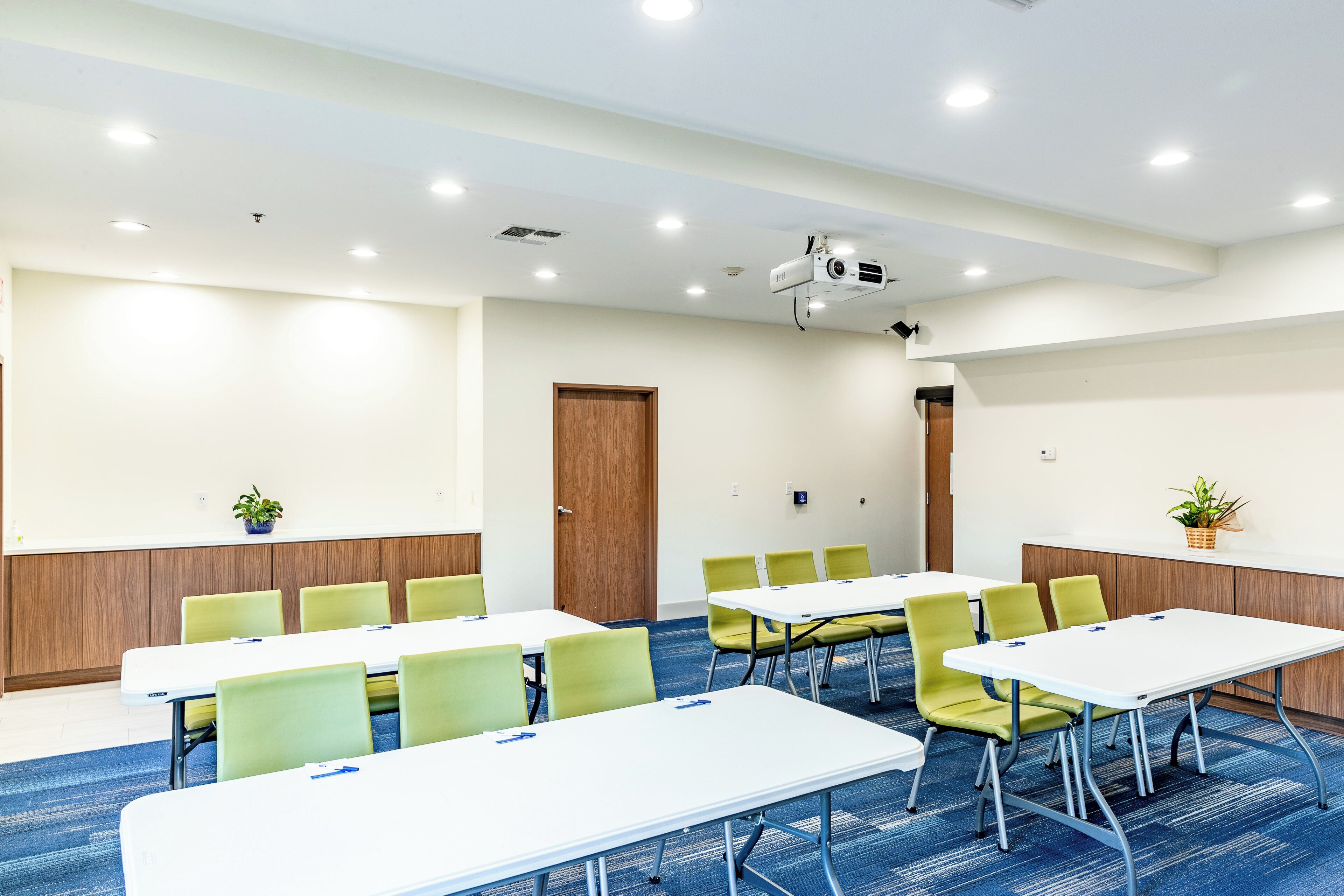 Uma sala de conferências com mesas brancas, cadeiras verdes, um projetor e um carpete azul.