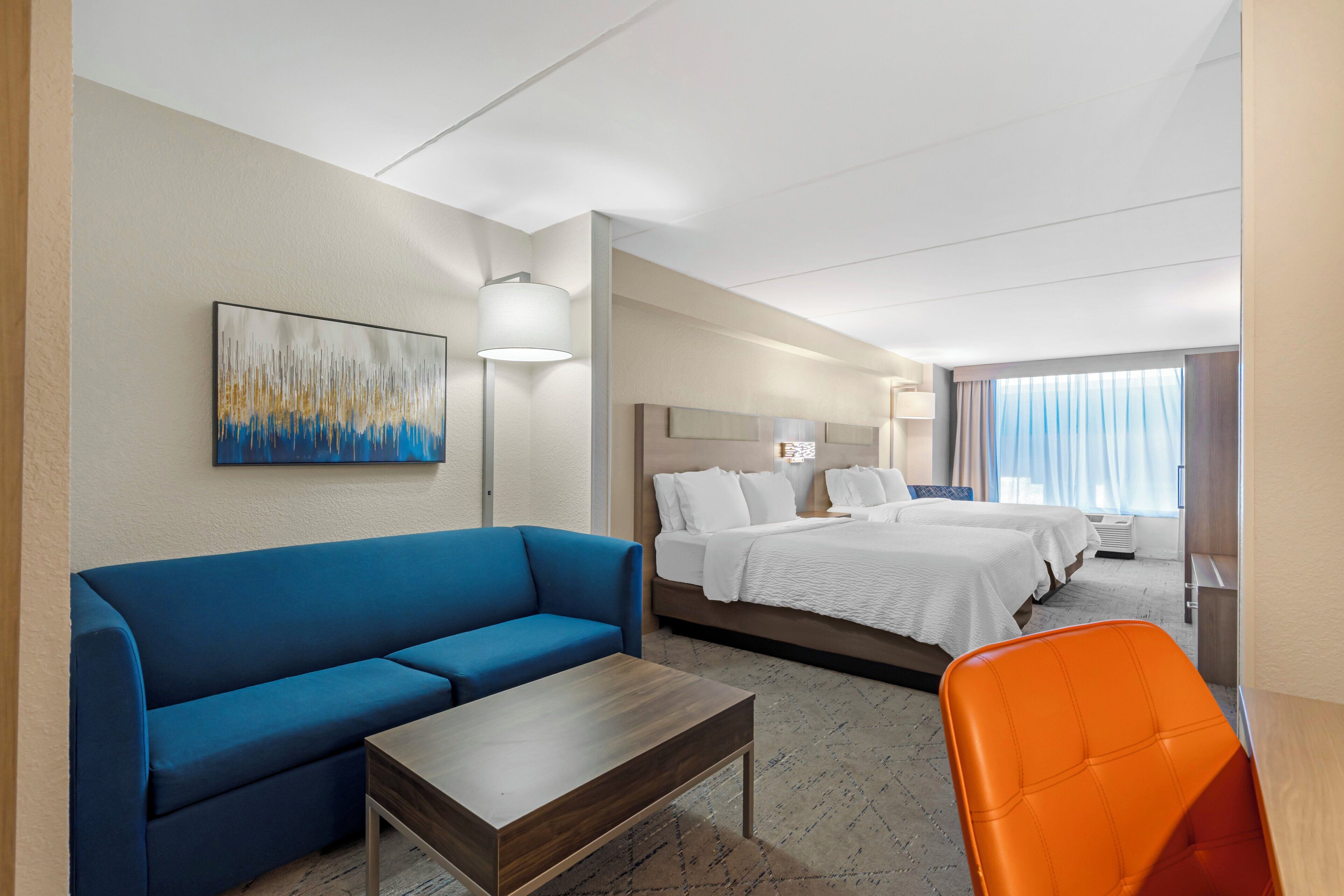 Habitación de hotel con un sofá azul, una mesa de centro de madera, una cama con ropa blanca y un sillón naranja brillante.