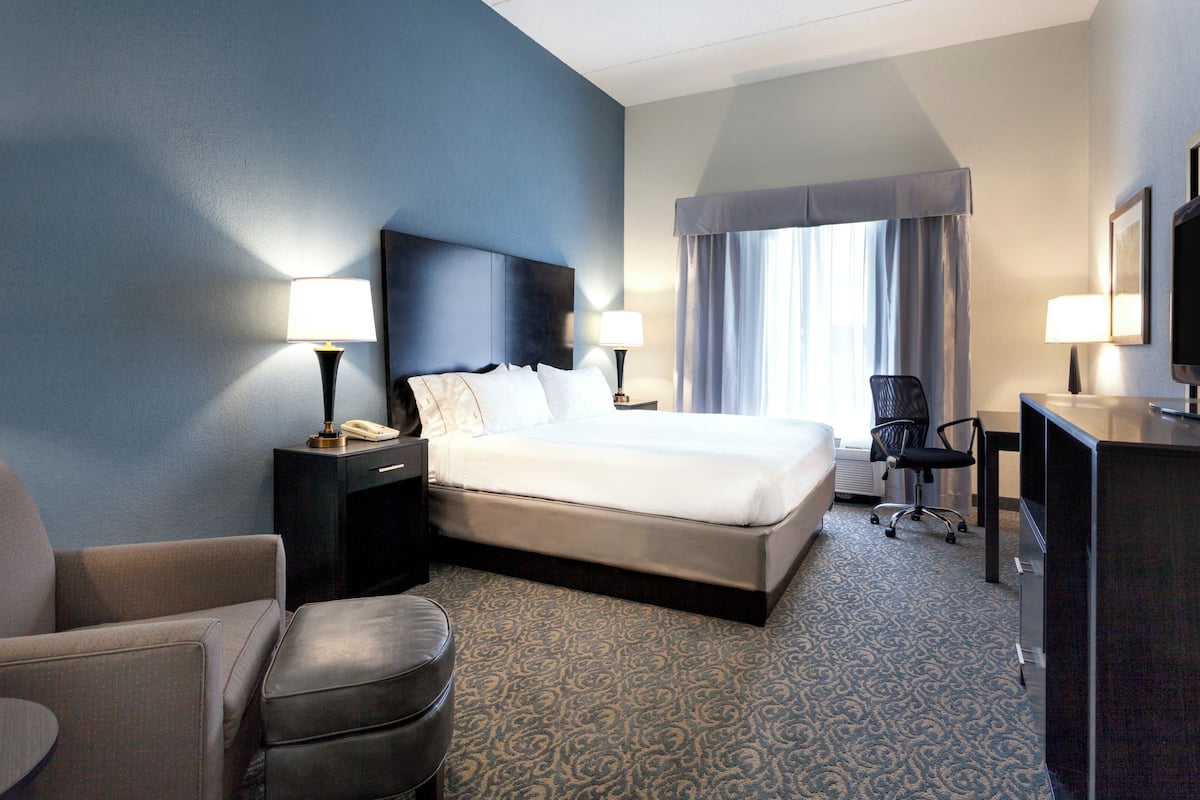 A hotel room with a large bed, two bedside lamps, a desk with a computer, a chair, and a small round table.
