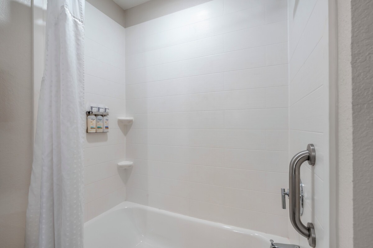 A bathroom with white tiled walls, a bathtub, a shower curtain, and a handrail.