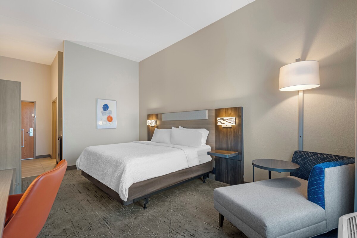 A hotel room with a bed, a chair, a small table, and a lamp.