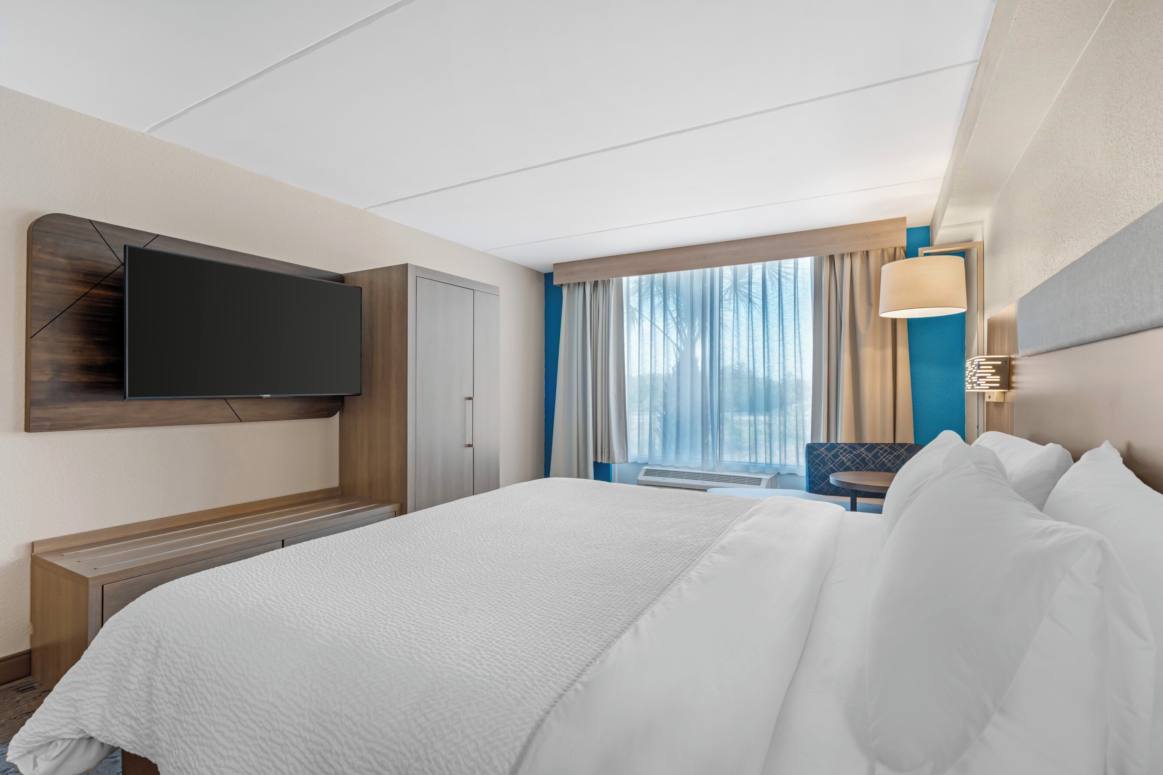 Habitación de hotel con cama, televisor montado en la pared, zona de estar con una mesa y ventana con cortinas.