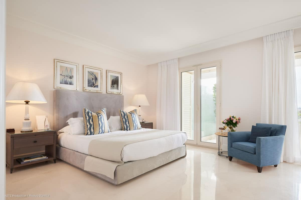 A modern bedroom with a large bed, two bedside tables, a blue armchair, and a view of the outdoors through a sliding door.