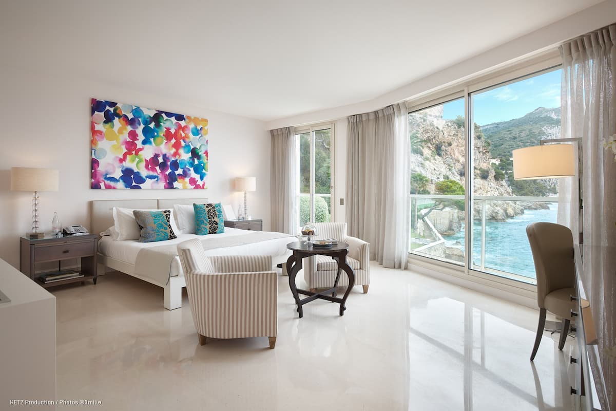 A modern hotel room with a large bed, two armchairs, a small table, and a view of the sea.