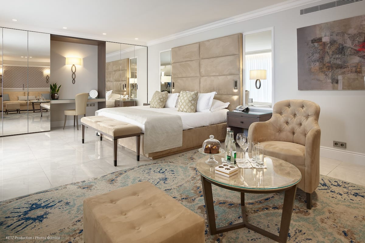 A modern hotel room with a large bed, a glass coffee table, a tufted armchair, and a dining area with a table and chairs.