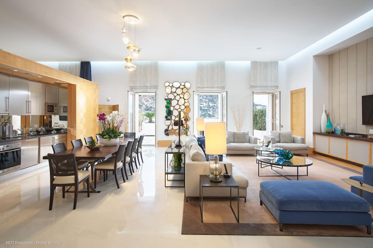 A modern open-plan living and dining area with a kitchen, a sofa, a coffee table, and a blue ottoman.