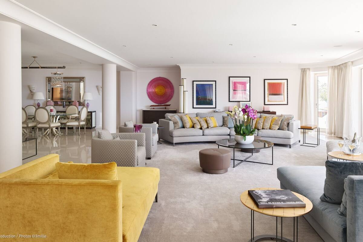 A spacious living room with a yellow sofa, grey armchairs, and a grey sofa with colorful cushions. There is a coffee table with a book and a vase of flowers. The room has a large window with curtains and a dining area in the background.