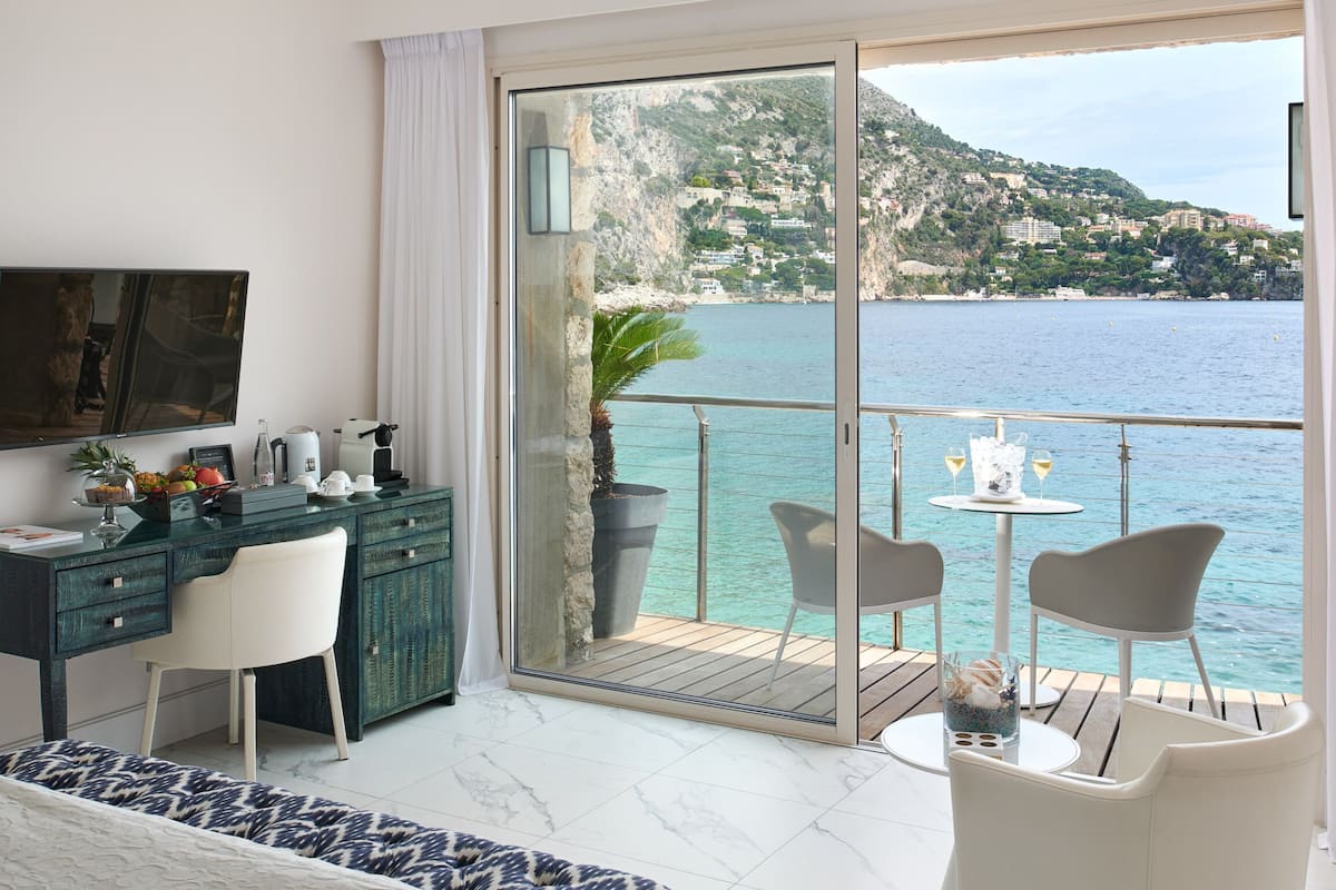 A bedroom with a bed, a desk, and a balcony with a view of the sea and a mountain.